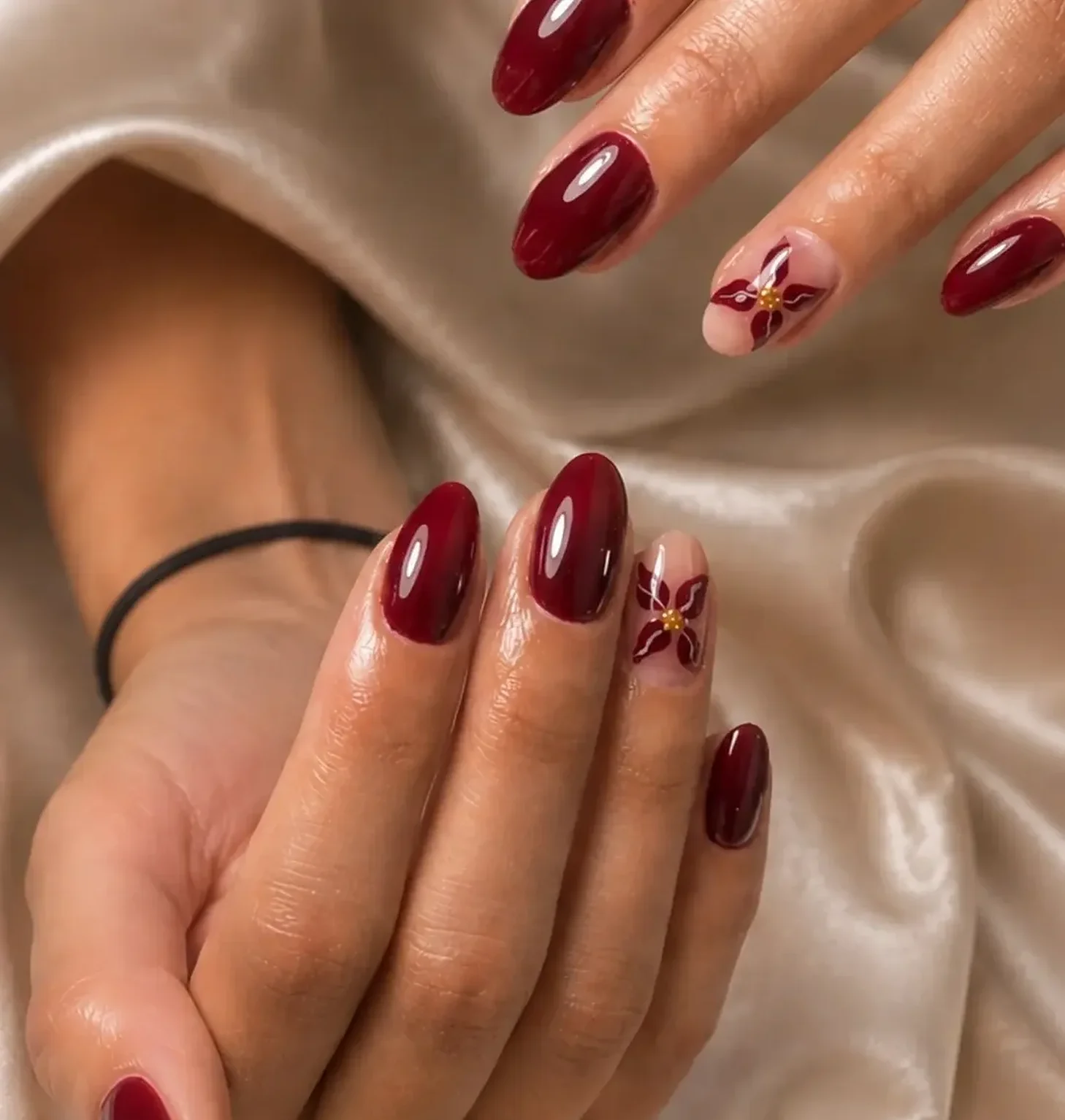 Glossy burgundy almond nails with a hand-painted flower accent on champagne satin